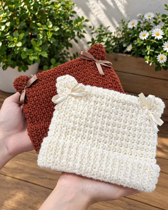 Dos gorritos de bebé tejidos al crochet, uno marrón y otro crudo, con orejitas y moños