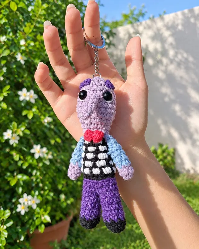 Llavero amigurumi de personaje lila con chaleco y moño rojo
