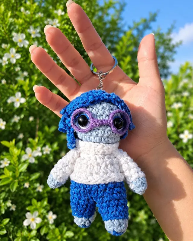 Llavero amigurumi de personaje azul con anteojos y buzo blanco